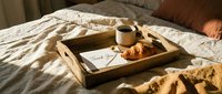 Wooden breakfast tray on bed with coffee, croissant, and thank you note in warm morning sunlight, cozy bedroom atmosphere.