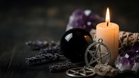 Mystical altar setup with burning candle, pentagram pendant, amethyst crystals, sage bundle and black scrying bowl on dark wooden surface for spiritual practices.