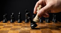 Hand moving white king chess piece on wooden board with black pieces in background during strategic game play.