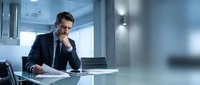 Confident businessman in suit sitting at modern office desk, hand on chin in thoughtful pose. Professional corporate executive planning strategy in contemporary workplace.