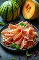 Sliced prosciutto ham with fresh cantaloupe melon and basil leaves on dark plate, gourmet appetizer for Mediterranean cuisine and fine dining.