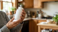 Senior woman holding blank white supplement bottle in modern kitchen, promoting health wellness and nutritional products for mature adults.