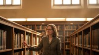 Professional blonde woman librarian browsing books in academic library with warm lighting and wooden shelves for educational content.