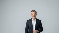Middle-aged man with gray hair and mustache wearing dark blazer and white shirt, standing against neutral background for corporate portraits and business headshots.