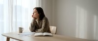Young Asian woman in cozy sweater reading book at wooden desk by window with coffee cup, peaceful morning study session in bright minimalist home office.