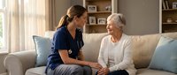 Young female healthcare worker in blue scrubs providing compassionate care to elderly Caucasian woman patient during home visit consultation in bright living room.