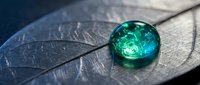 Emerald green water droplet resting on textured leaf surface with detailed vein patterns, macro photography showcasing nature's intricate beauty and freshness.