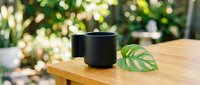 Black ceramic mug on wooden table with green leaf in natural outdoor setting with bokeh background for lifestyle and home decor concepts.