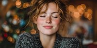 Young Caucasian woman with curly blonde hair and closed eyes wearing cozy gray sweater enjoying peaceful moment with warm bokeh lights background.
