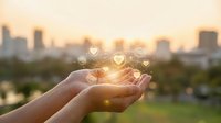 Hands cupping glowing heart icons with warm sunlight and blurred city skyline background representing love, care, charity and human connection concepts.