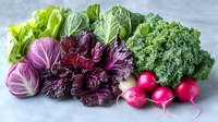 Fresh organic vegetables arranged on gray surface including purple cabbage, curly kale, crisp lettuce, and bright red radishes showing vibrant natural colors and textures.
