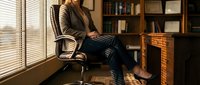 Confident professional woman sitting in leather office chair by window, warm light, bookshelf background, business casual attire, corporate setting.