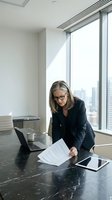 Middle-aged woman with gray hair and glasses wearing black blazer reviewing documents at modern office desk with laptop and tablet, city skyline visible through floor-to-ceiling windows.