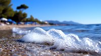 Sea foam bubbles on sandy beach shore with gentle waves, crystal clear water and blurred mountain silhouette in background on sunny summer day.