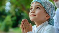 Young boy in traditional Islamic headwear praying outdoors with hopeful expression, looking up at sky against blurred green nature background, expressing faith and spirituality.