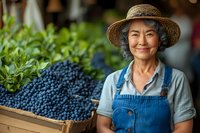 Senior Asian woman farmer in straw hat and denim overalls smiling warmly next to wooden crates filled with fresh harvested blueberries at local farm market.