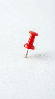 Red push pin thumbtack stuck in white textured surface for office organization and bulletin board displays.