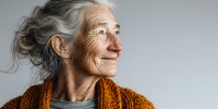 Elderly woman with gray hair and wrinkles smiling gently while wearing an orange knitted scarf, representing aging with grace and wisdom.