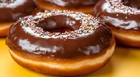 Chocolate glazed donut with colorful sprinkles on yellow background, close-up view showing glossy texture and decorative toppings. Fresh baked pastry.