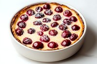 Fresh baked cherry clafoutis in white ceramic baking dish, traditional French dessert with whole cherries and custard dusted with powdered sugar.