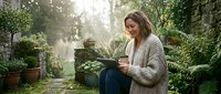 Young woman with light brown hair and fair complexion working on laptop in lush garden surrounded by green plants and ivy-covered walls, natural sunlight filtering through foliage.