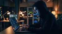 Young Asian male programmer coding on laptop in dark office with city lights background, focused developer working late night on software project.