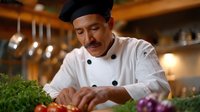 Hispanic male chef in traditional white uniform and black hat carefully preparing fresh herbs in upscale restaurant kitchen with warm ambient lighting.
