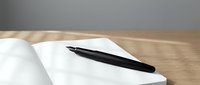Black pen resting on open blank notebook with white pages on wooden desk surface against gray wall background for writing and note-taking.