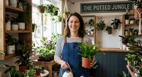 Smiling young woman in striped shirt and denim overalls holding potted plant in cozy indoor garden shop filled with houseplants and greenery.