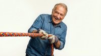 Senior Caucasian man in denim shirt pulling colorful rope in tug of war game, showing determination and strength in competitive activity on white background.