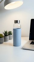 Glass water bottle with blue silicone sleeve on modern white desk next to laptop and succulent plants under warm pendant light for healthy hydration lifestyle.