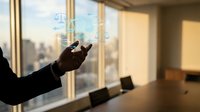 Businessperson in dark suit interacting with holographic digital interface displaying connected network symbols above hand, modern office with city skyline view through windows.