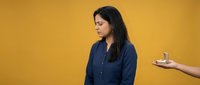 Young Hispanic businesswoman in navy shirt looking at smartphone offered by hand against vibrant yellow background for corporate communication concepts.