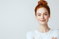 Young woman with red hair and natural makeup smiling confidently with arms crossed against light background, expressing positive professional attitude.