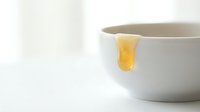 Honey dripping from wooden spoon into white ceramic bowl on bright background, isolated for cooking and baking design projects.