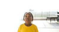Young Black child wearing bright yellow shirt looking upward with thoughtful expression in modern interior space with natural light.
