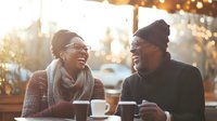 Young African American couple laughing together at outdoor cafe during golden hour, sharing coffee and conversation with warm bokeh lights in background.
