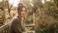 Young woman with dark hair in cozy brown sweater sitting outdoors during golden hour autumn light with warm bokeh background for lifestyle content.