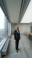Middle-aged Caucasian businesswoman in dark blazer standing in modern office corridor with floor-to-ceiling windows, concrete flooring, and minimalist interior design for corporate, professional, and business concepts.