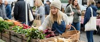 Young blonde woman in beige knit hat shopping for fresh vegetables at outdoor farmers market with wicker basket, selecting organic produce from wooden crates.