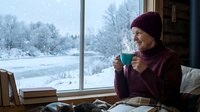Young Caucasian woman in burgundy sweater and beanie enjoying hot drink by window overlooking snowy winter landscape from cozy indoor setting.