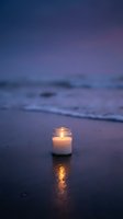 Glowing candle in glass jar on wet beach sand at twilight with ocean waves and purple sky creating peaceful meditation atmosphere for relaxation concepts.