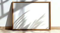 Empty wooden picture frame on white wall with natural shadow overlay from window light creating palm leaf silhouette effect, minimal home interior design element.