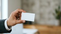 Businessman holding blank white business card in office environment with blurred background for corporate branding and professional networking purposes.