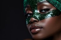 Close up portrait of African American woman wearing sparkling emerald green carnival mask with glossy lips and dramatic eye makeup on dark background.