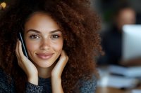 Young African American woman with natural curly hair smiling warmly while holding face in hands. Soft natural lighting highlights genuine expression in professional office setting.