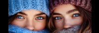 Two young Caucasian women wearing knitted winter hats and scarves covering lower faces, showing bright blue eyes and warm smiles for cold weather fashion.