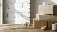 Stacked kraft paper gift boxes with twine bows on wooden table against white brick wall, decorated with dried flowers and white mug for holiday packaging and retail display.