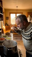 Senior Caucasian man with gray beard cooking soup in warm kitchen wearing patterned sweater tasting homemade meal from wooden spoon during golden hour.