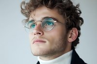 Young caucasian male with curly hair wearing round metal glasses and turtleneck sweater looking up thoughtfully against light background. Professional portrait in soft lighting.